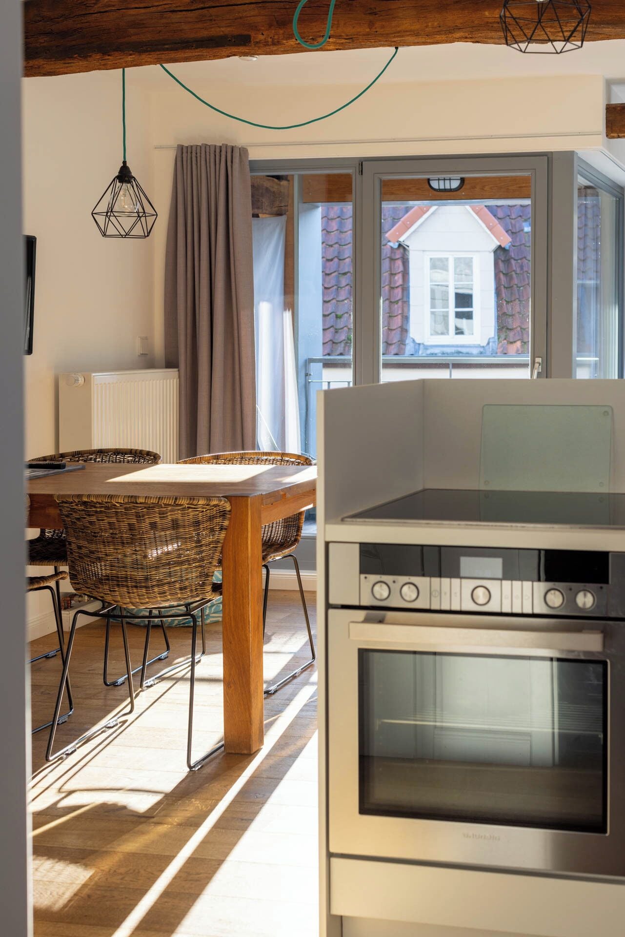 A kitchen with a built-in oven, a dining table with wicker chairs, and a view of a house through a window.