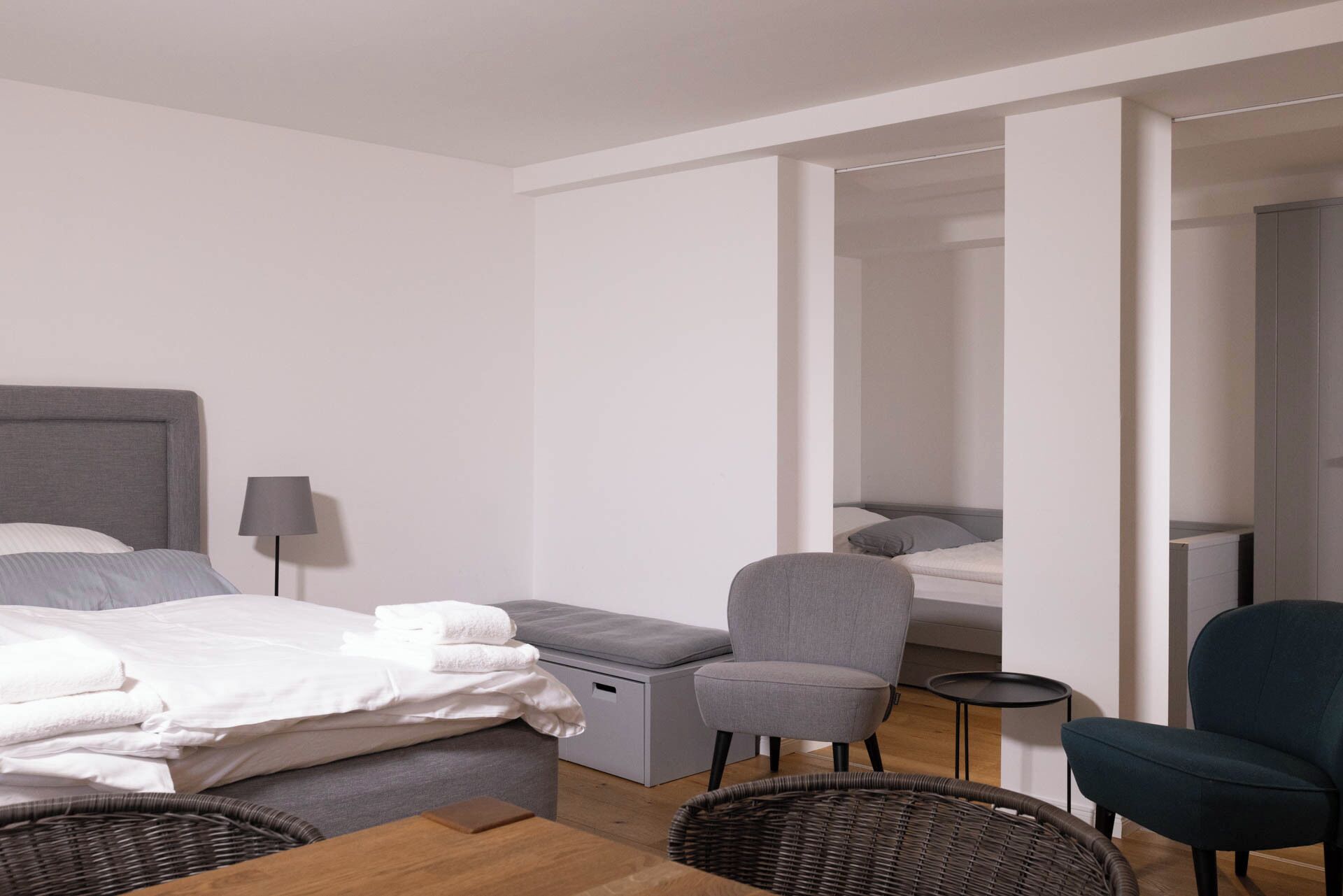 Moderni hotellihuone, jossa on sänky, harmaa nojatuoli, pieni pyöreä pöytä ja rottinkituoli.