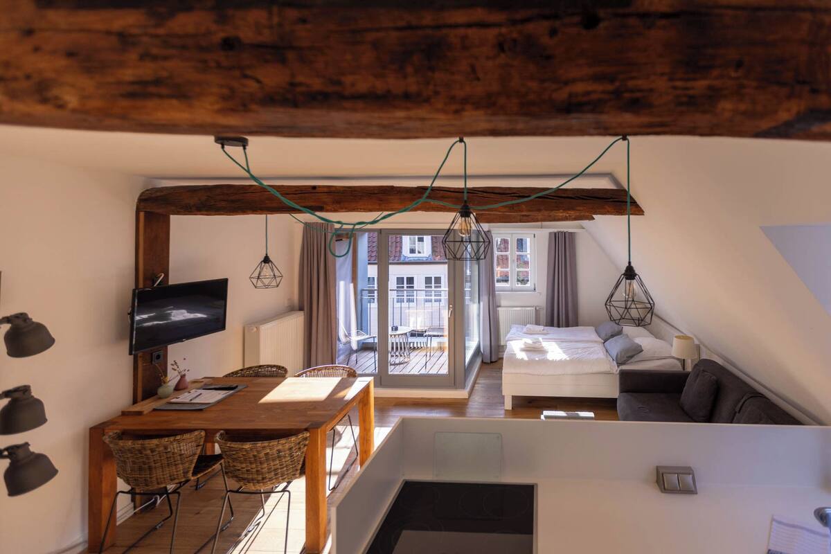 A cozy room with a wooden ceiling, a dining area with wicker chairs, a bed with a gray sofa, and a TV on the wall.