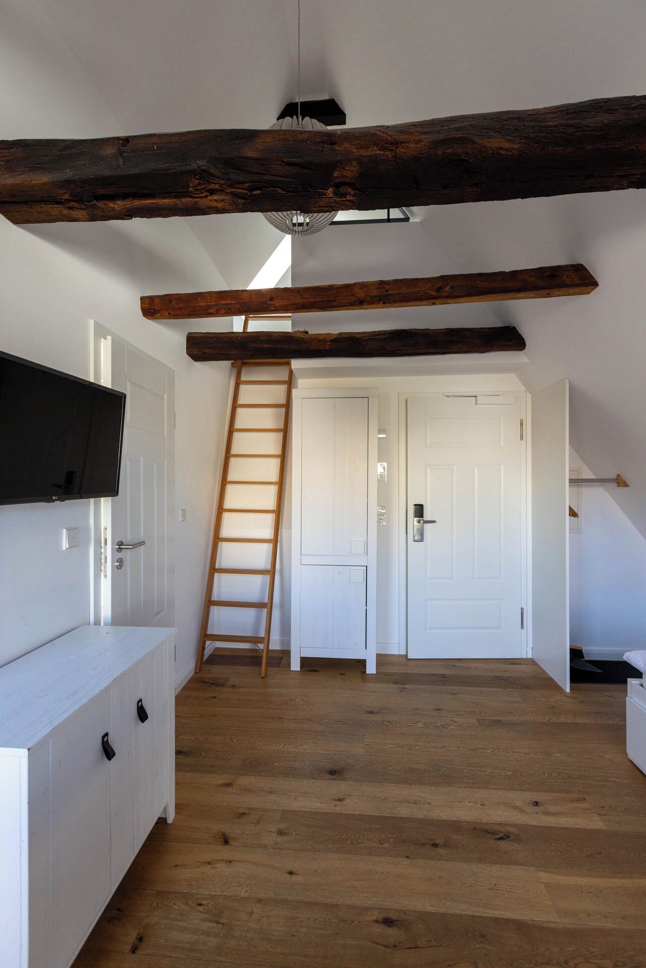 A spacious attic room with exposed wooden beams, a wooden ladder, white doors, and a white cabinet.