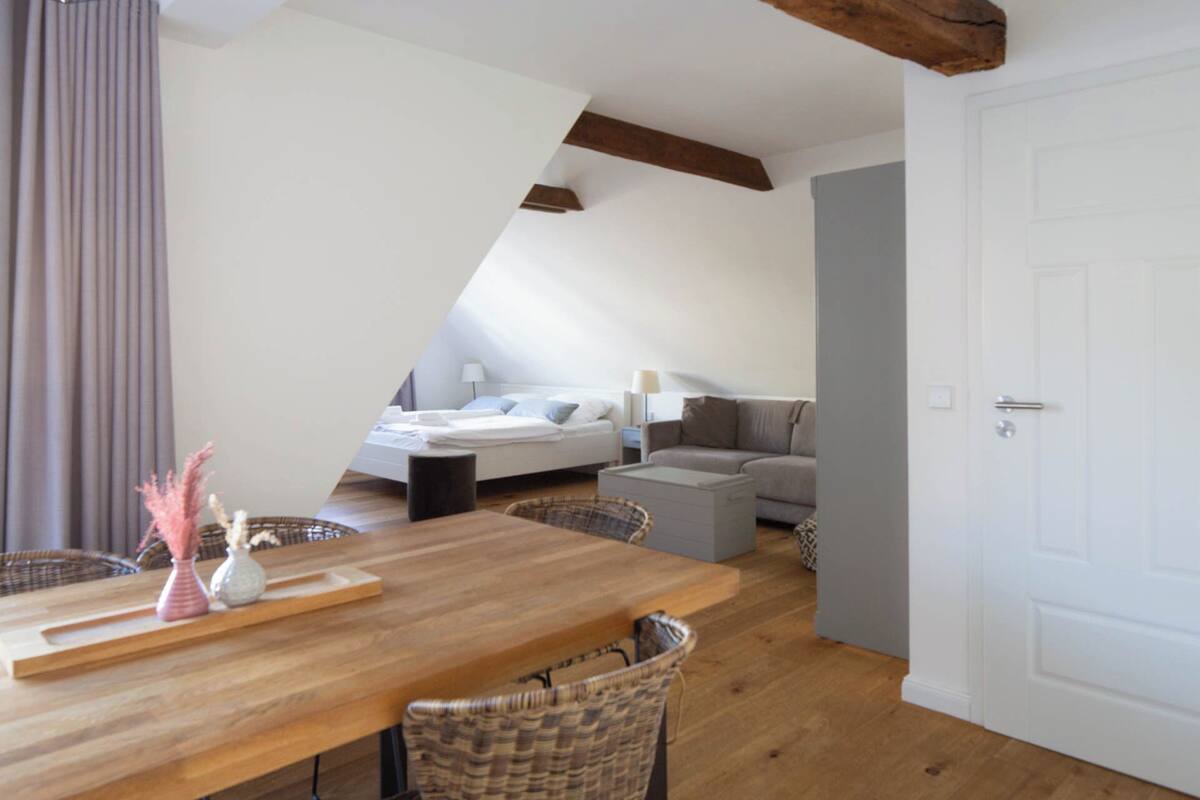 A modern loft bedroom with a sloped ceiling, a bed, a sofa, and a dining area with a wooden table and wicker chairs.