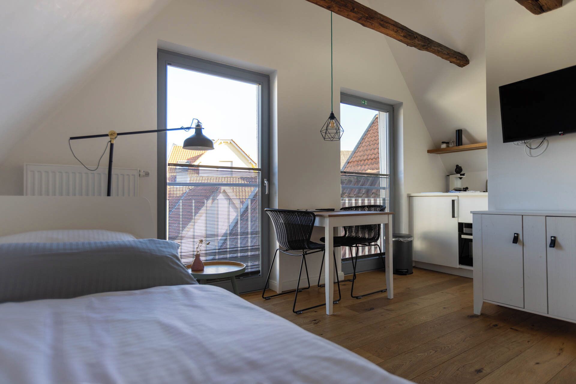 A bedroom with a bed, a desk, and a chair. There is a window, a radiator, and a television on the wall.