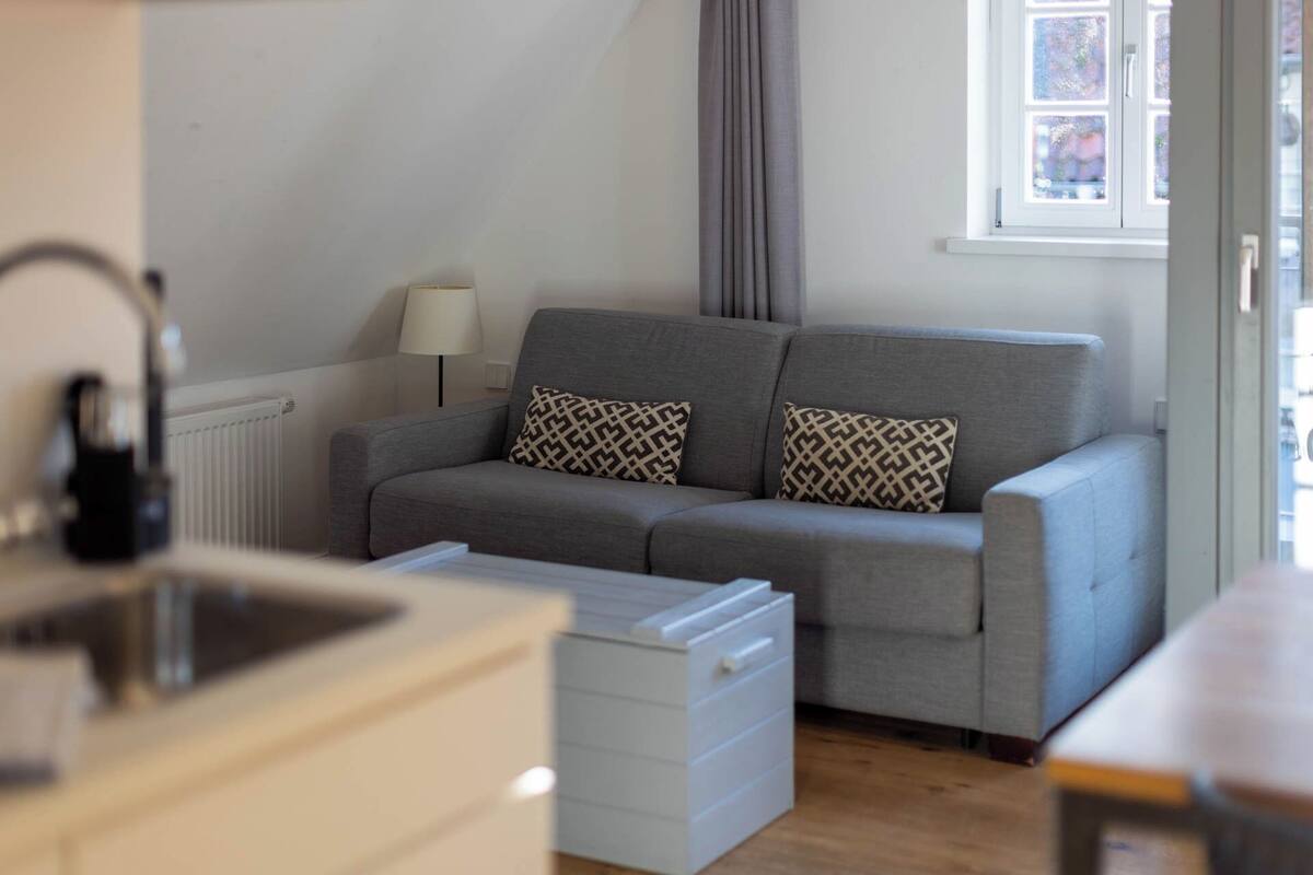 A modern living room with a grey sofa, patterned cushions, a white side table, and a window with a view.