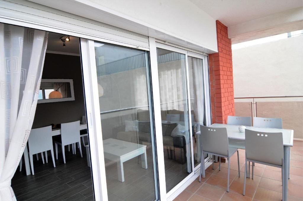 A balcony with a dining area, including a table and chairs, and a sliding glass door leading to an indoor space with a sofa and a lamp.