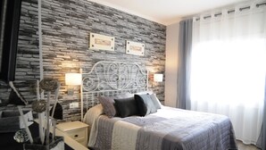 A bedroom with a stone wall, a bed with a metal headboard, bedside lamps, and a window with curtains. - HomeHolidaysRentals  Maritim V (Pineda de Mar)