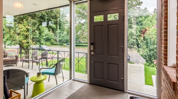 A room with a large glass door leading to an outdoor patio area with wicker furniture and a view of a street and greenery.