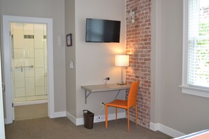 Habitación con ducha, escritorio con silla, televisor montado en la pared y ventana con persianas.