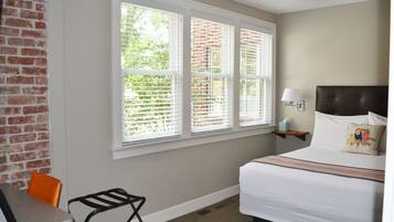 A hotel room with a bed, a desk, and a chair. There is a window with blinds, a nightstand with a lamp, and a brick wall.
