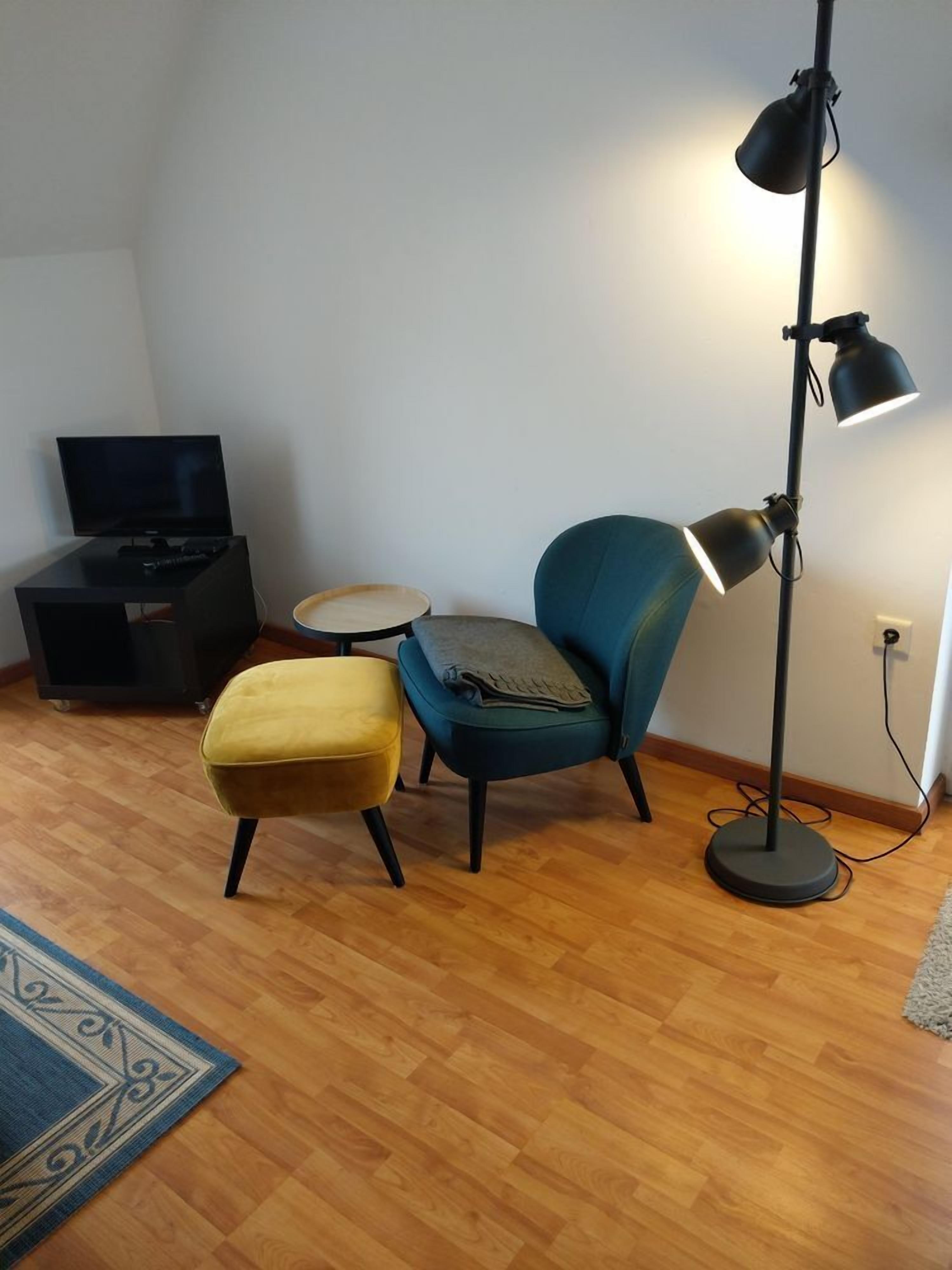 A living room with a television, a blue armchair, a yellow ottoman, a small round table, and a floor lamp.