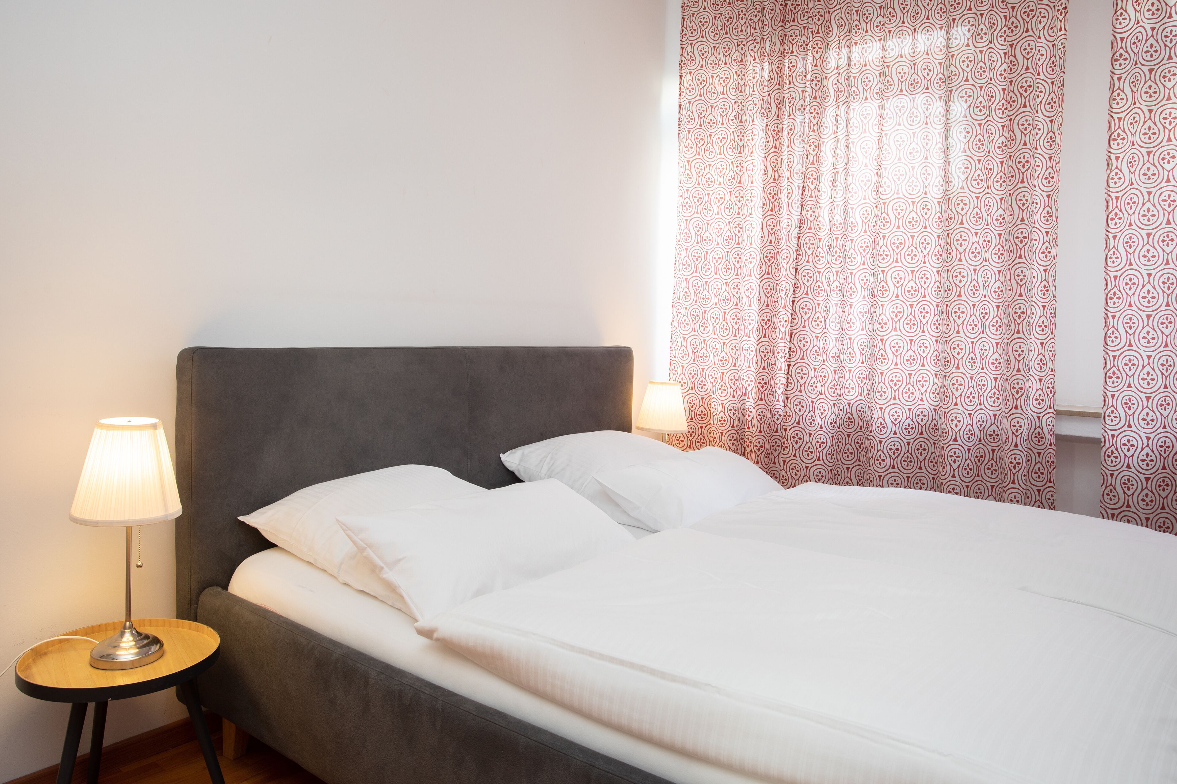 Una cama bien hecha con ropa blanca, un cabecero tapizado en gris y una mesita de noche con una lámpara.
