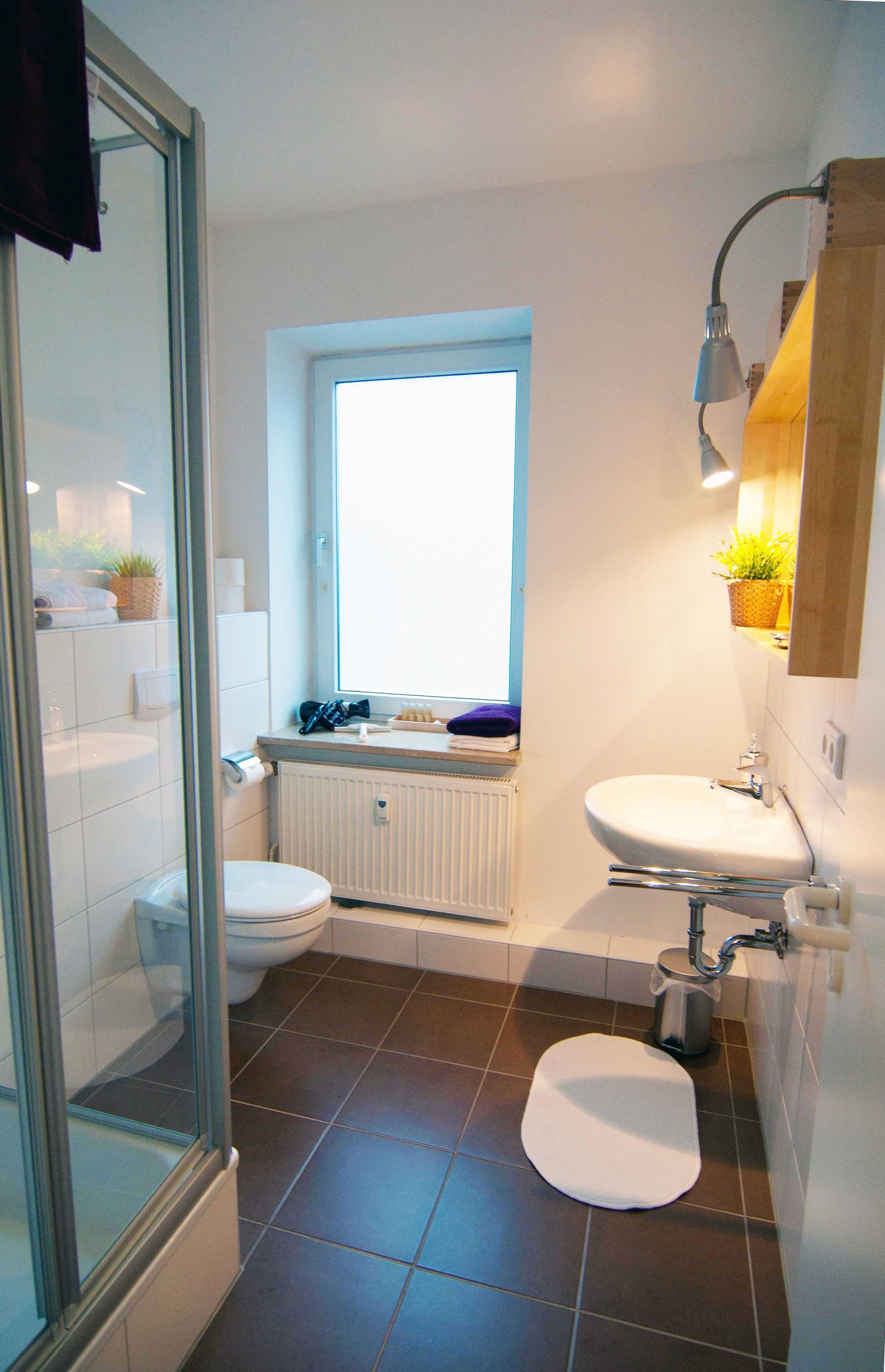 Ein Badezimmer mit Toilette, Waschbecken und Dusche. Über der Toilette befindet sich ein Fenster, und auf dem Fensterbrett steht eine Pflanze.