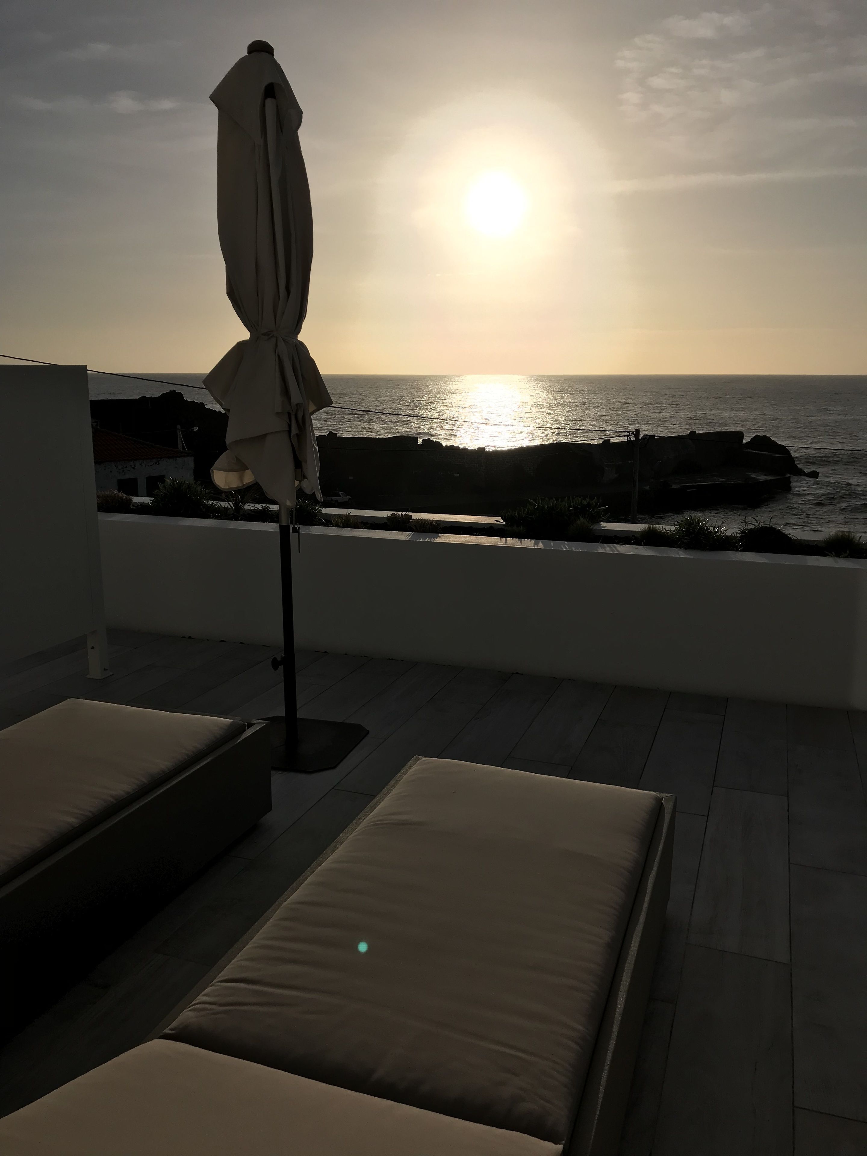 A balcony with a view of the sea at sunset, featuring two cushioned seating areas and a sun umbrella.