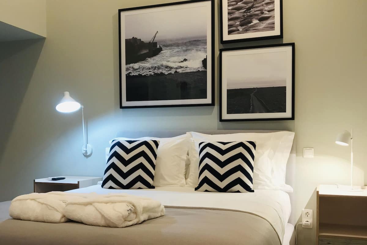 A neatly made bed with a beige blanket and two pillows, a bedside lamp, and a framed photograph of a coastal scene on the wall.