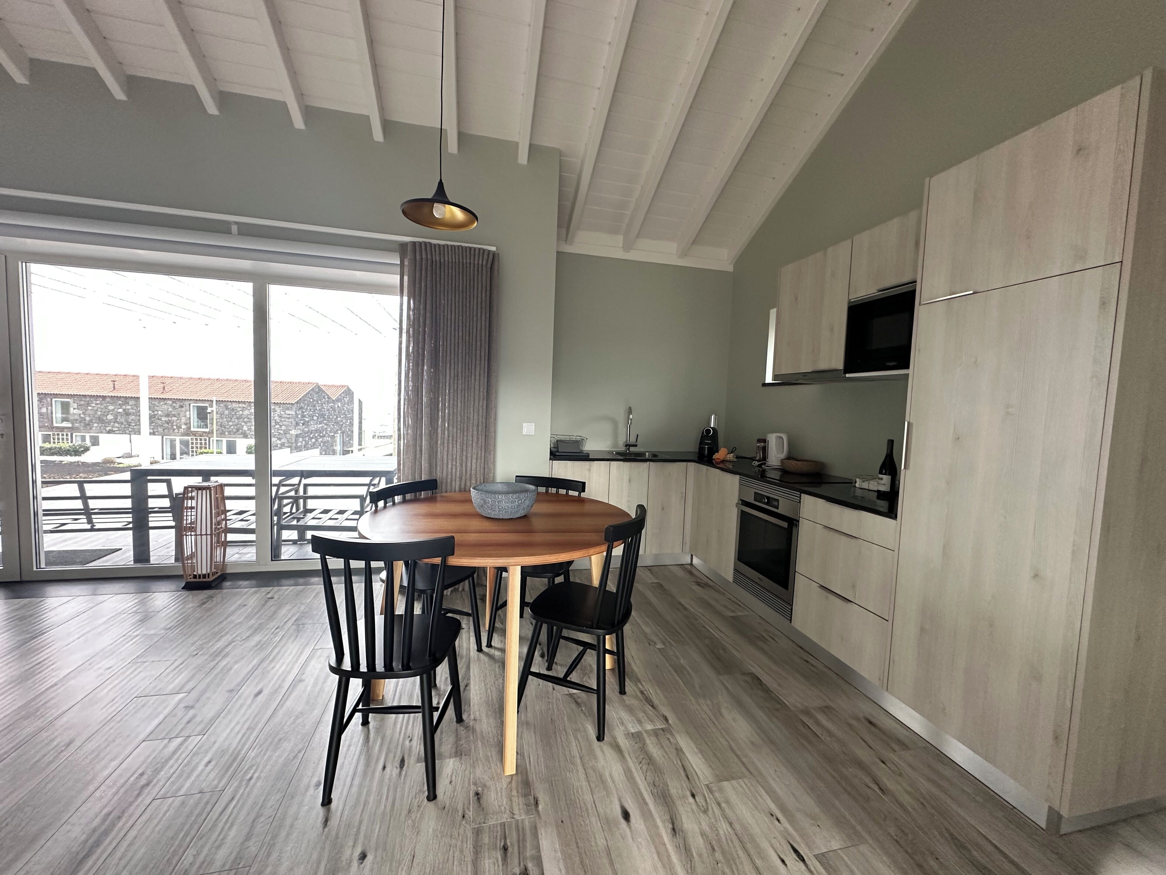 Eine moderne Küche mit Holzausstattung, einem Esstisch mit Stühlen und einem großen Fenster mit Blick auf eine Terrasse.