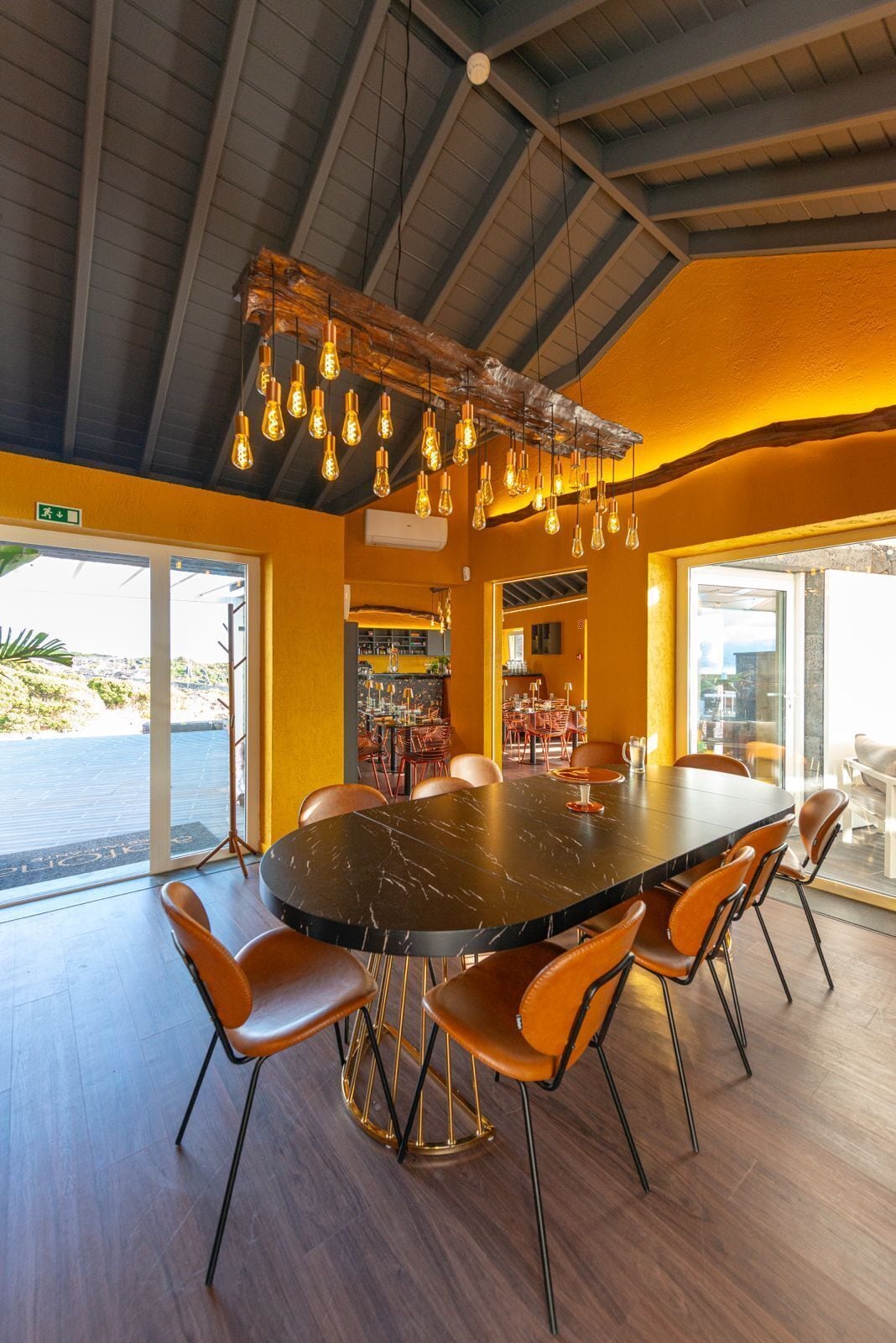 Un comedor espacioso con una gran mesa de mármol negro rodeada de sillas color naranja, una lámpara moderna y ventanas amplias que ofrecen vistas al exterior.