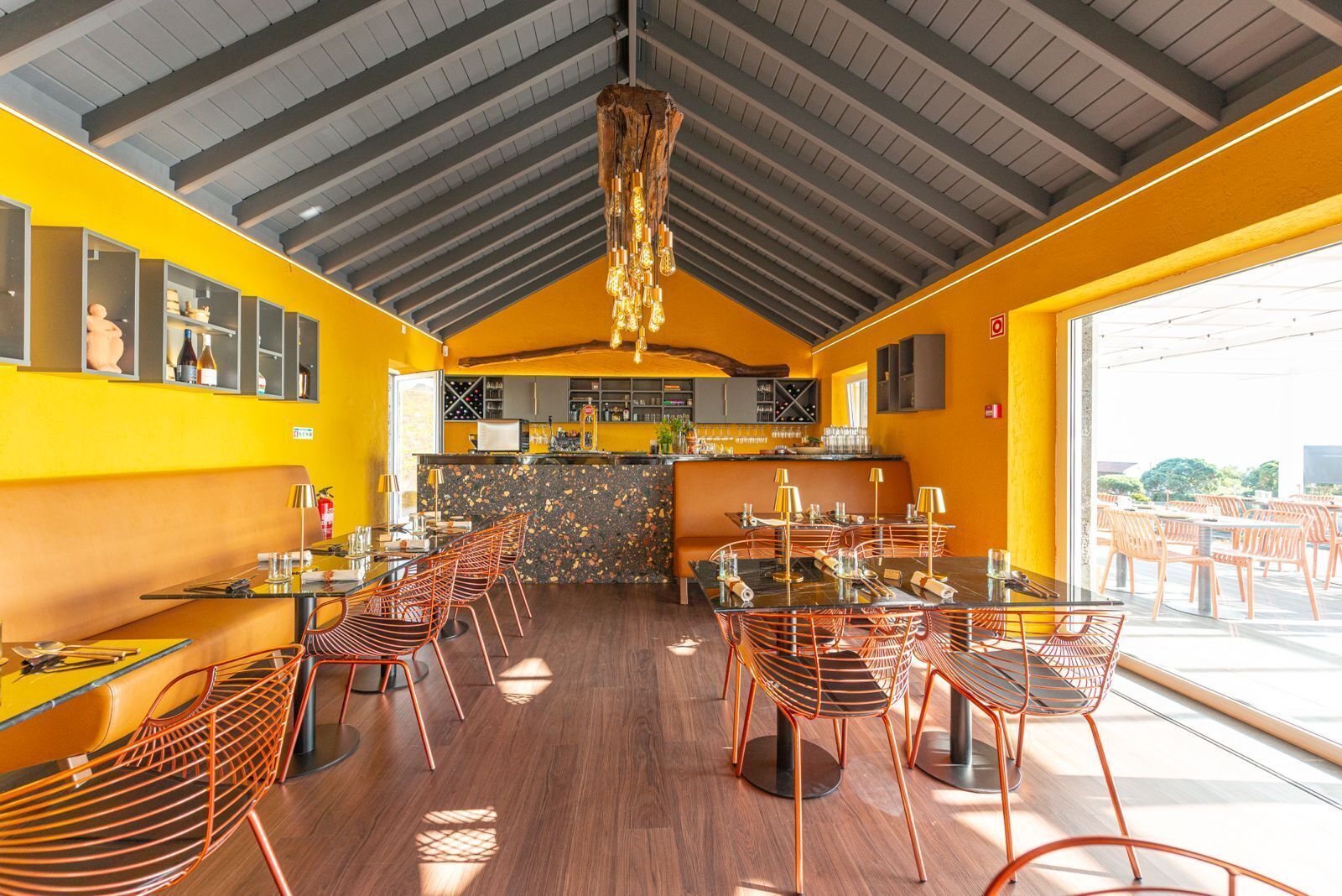 Un restaurant avec un plafond haut, des poutres en bois et un intérieur jaune vif. Les tables et les chaises sont disposées pour accueillir les clients.