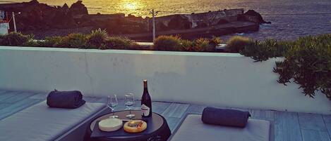 En tagterrasse med havudsigt i solnedgang, et bord med vinglas og en flaske, samt komfortable loungeområder.