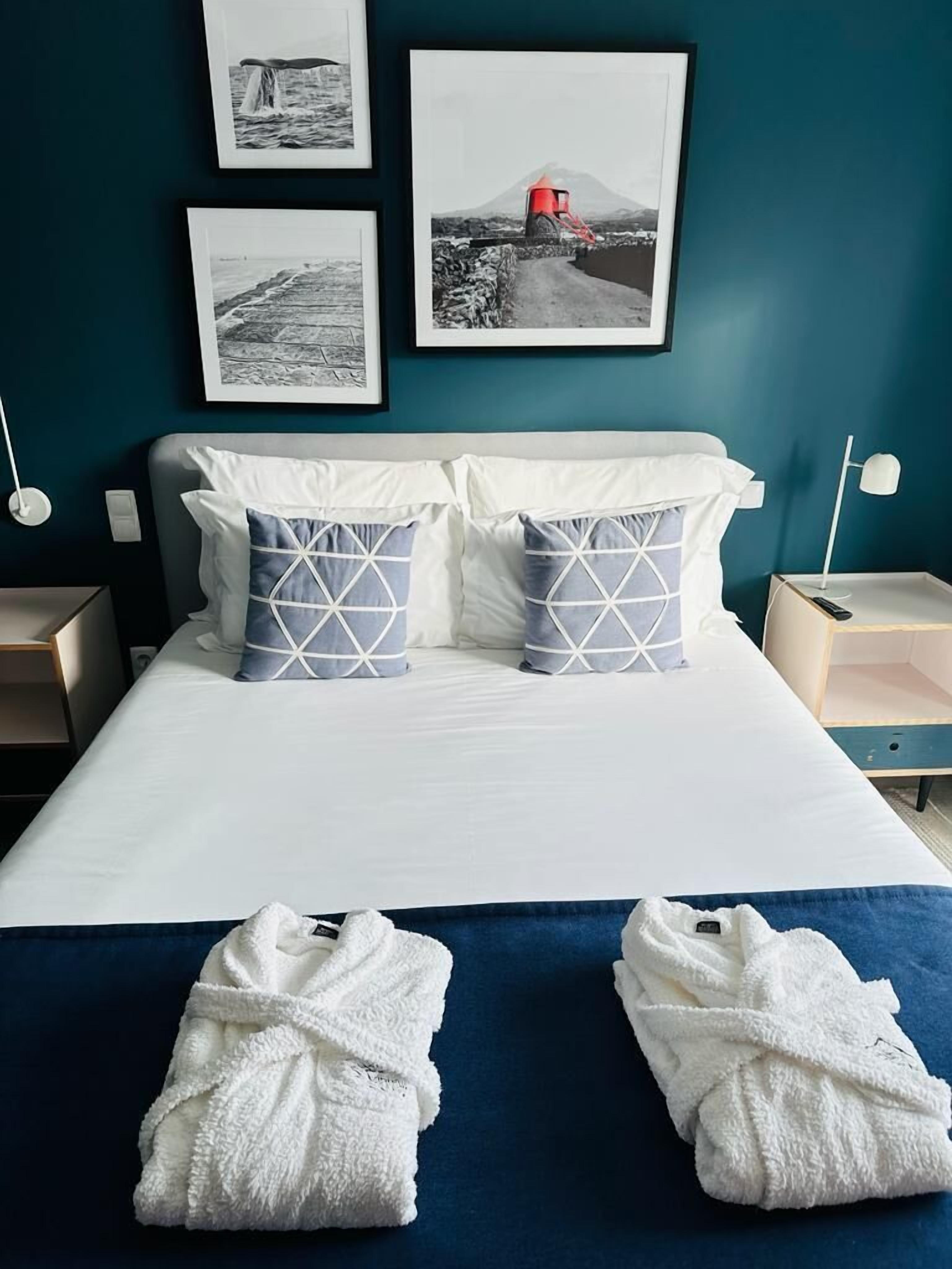 Una cama bien hecha con sábanas blancas, dos almohadas con diseños geométricos y dos toallas blancas sobre un edredón azul.