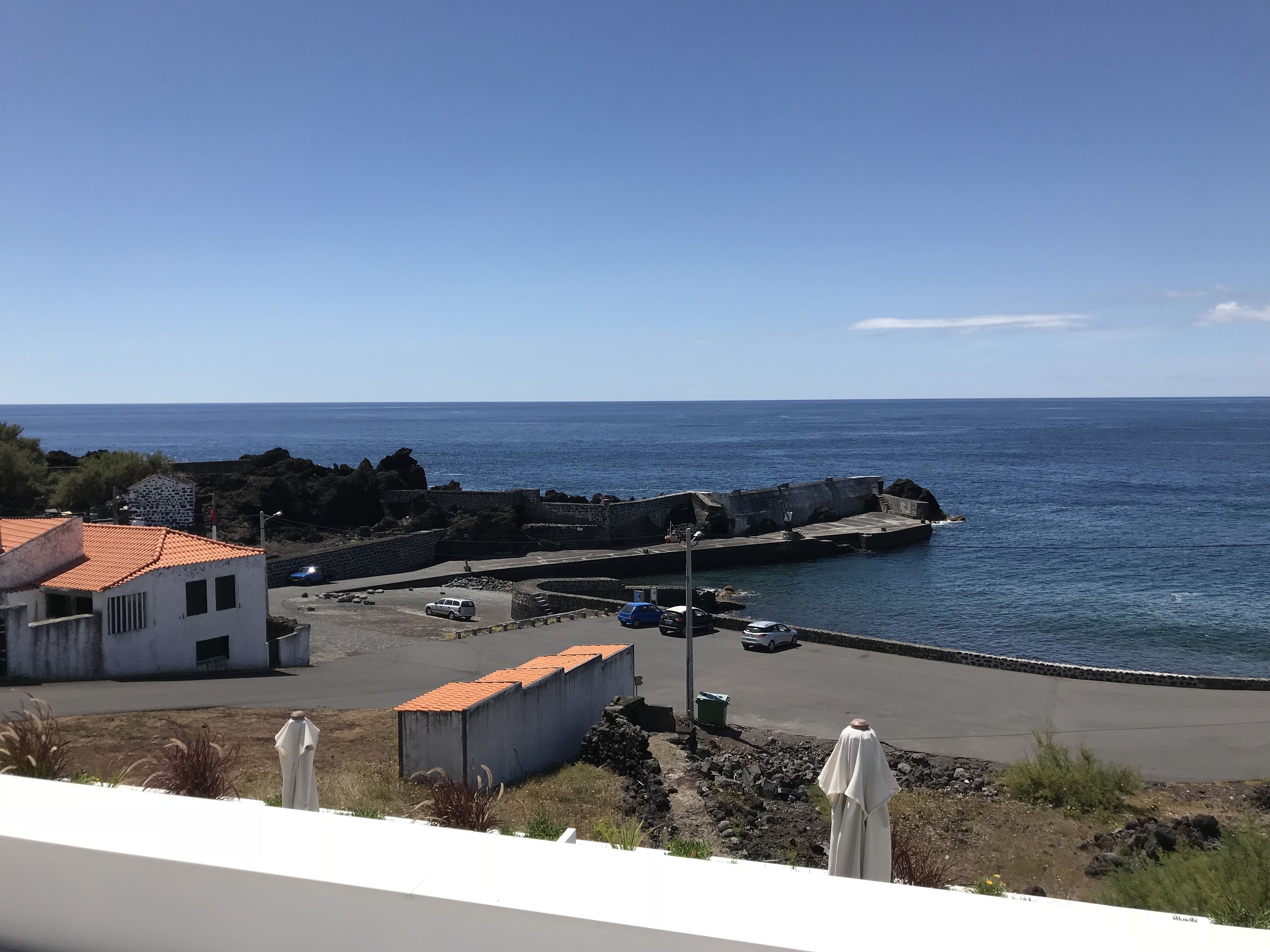 Vista costera con muelle de piedra, coches aparcados y un edificio con tejado de tejas rojas.