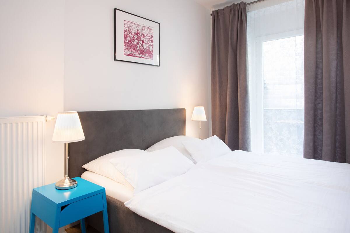 Quarto de hotel com cama, duas luminárias de cabeceira, um quadro na parede e janela com cortinas.