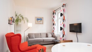 A modern living room with a grey sofa, a red armchair, a round white table, a flat-screen TV, and a window with polka dot curtains. - Wertschätzer II - Unter der Wehme (Detmold)