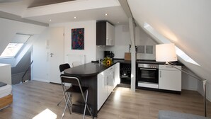 A modern kitchen with a central island, a dining area with a chair, and a staircase leading to an upper level. - Wertschätzer II - Unter der Wehme (Detmold)