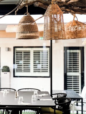 Wicker hanging lights, a table with chairs, and a window with blinds. - Hôtel Restaurant Le Nelio (Aix-en-Provence)