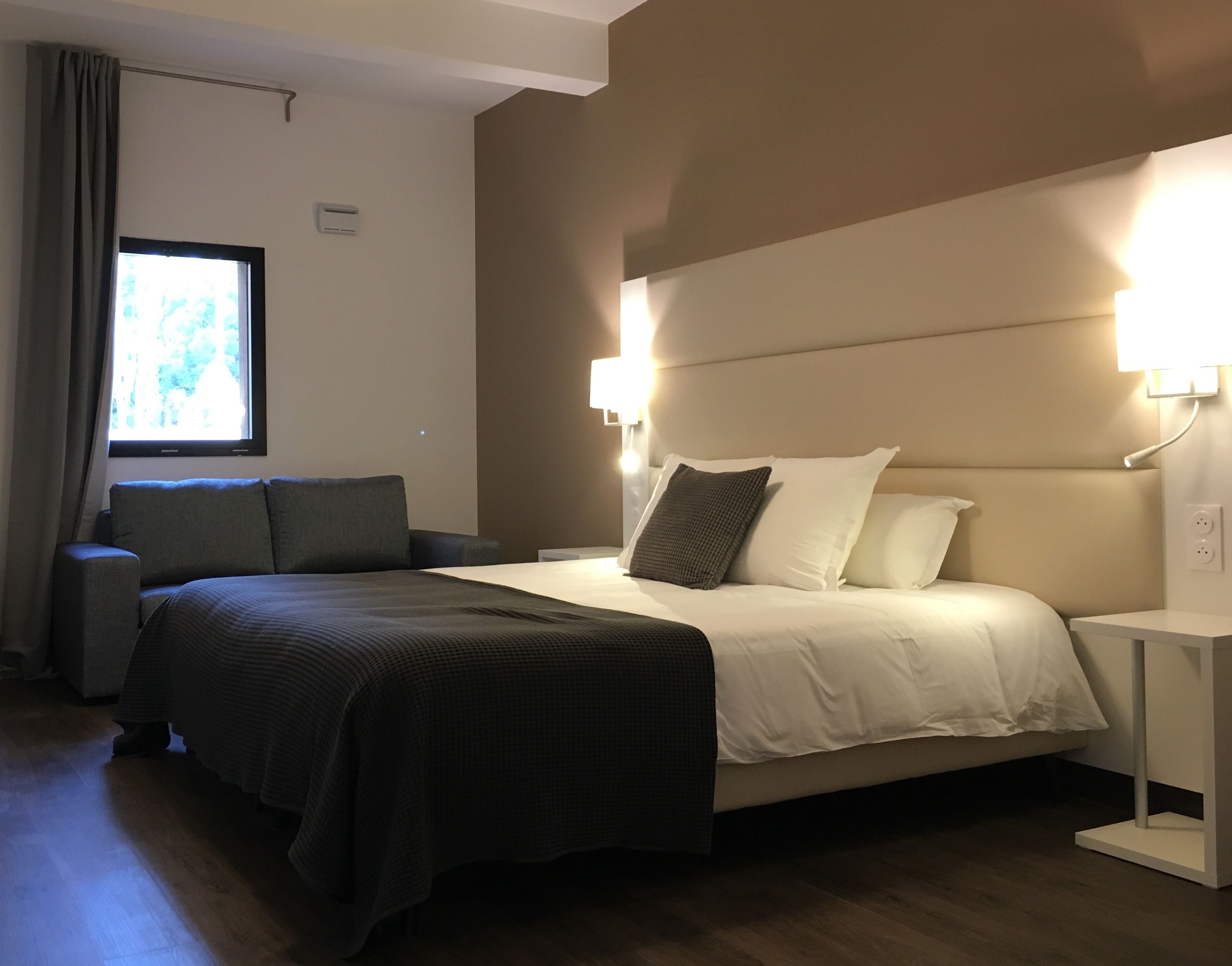 A hotel room with a bed, bedside tables, a window with curtains, and a wall-mounted light fixture.
