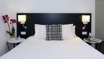 A modern hotel room with a large bed, two bedside lamps, a small table with a book and a bottle, and a potted plant.