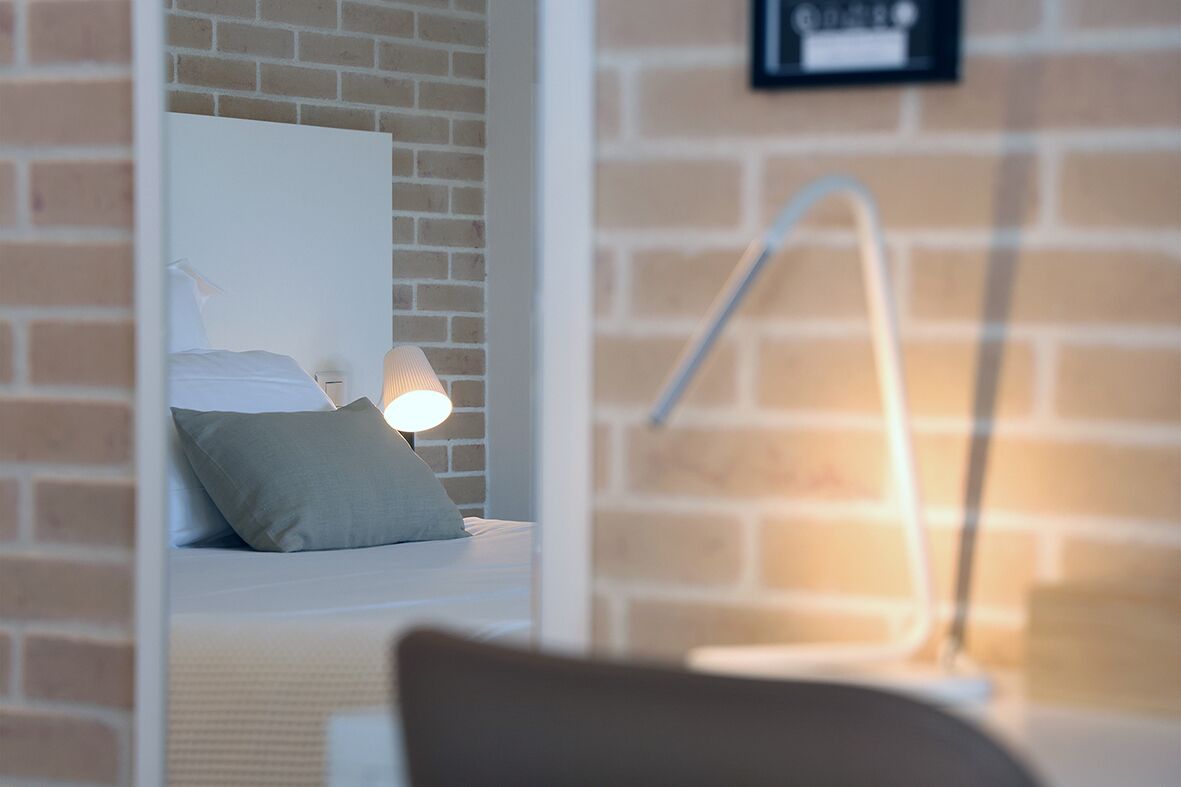 Ein modernes Zimmer mit Backsteinwand, einem Bett mit weißer Bettwäsche, einem Nachttisch mit Lampe und einer sichtbaren Badezimmertür.