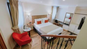 A hotel room with a bed, a red chair, a small table with a lamp, and a mirror on the wall.