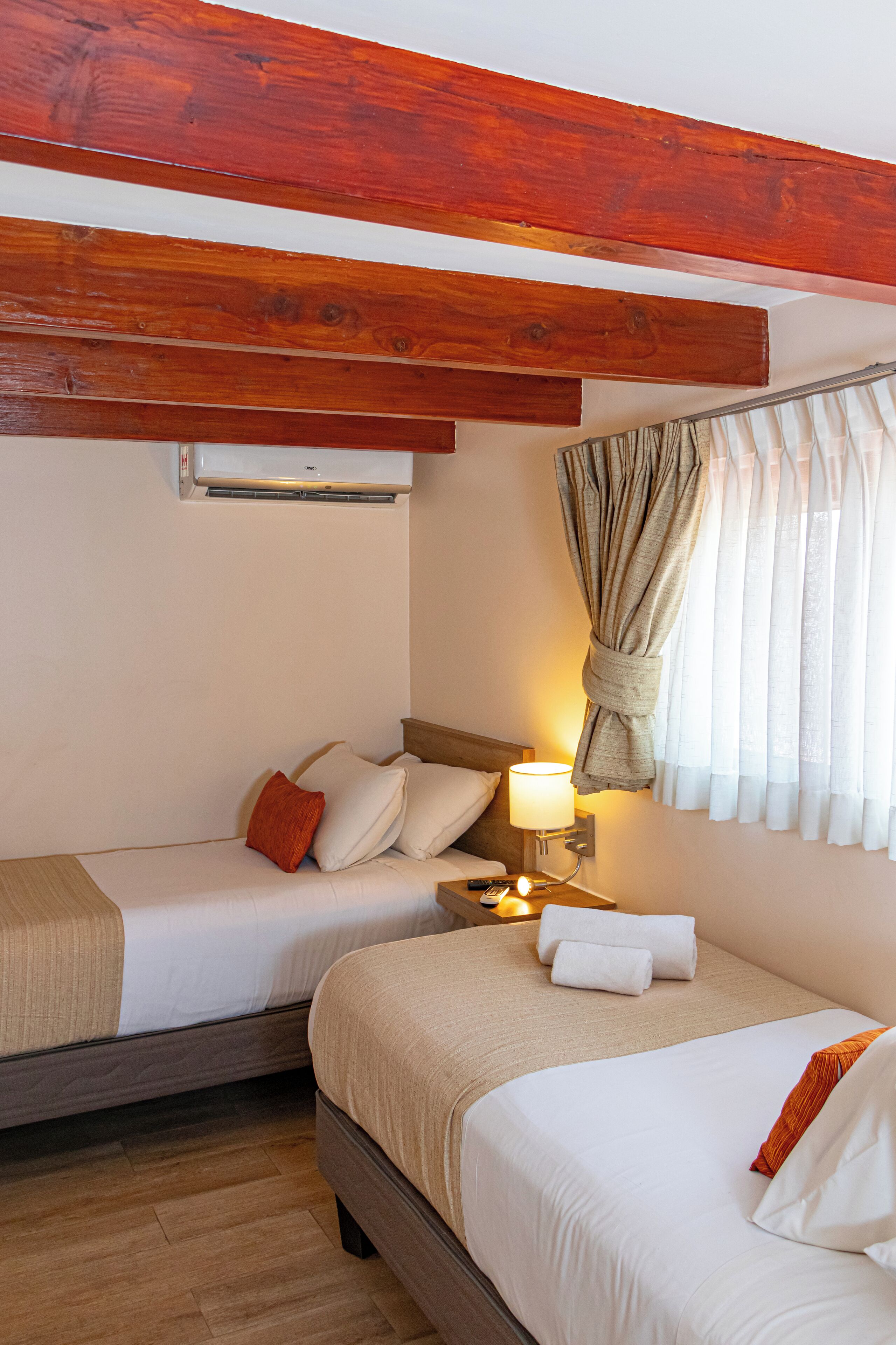 A hotel room with two beds, wooden beams, a wall-mounted air conditioner, a bedside lamp, and a curtain.