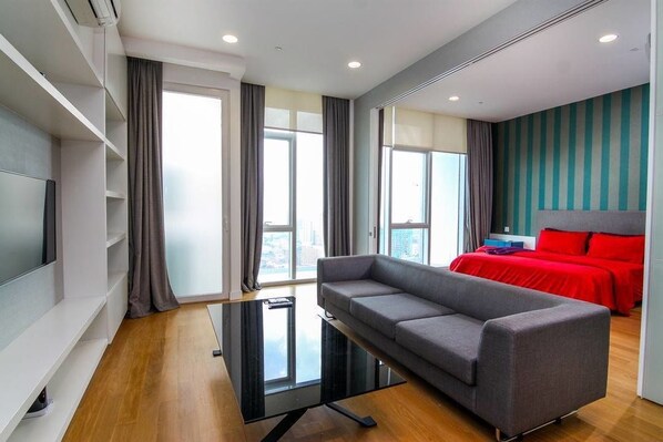 A modern hotel room with a grey sofa, a red bed, and a large window offering a city view. - Two Bedrooms Platinum Residence (Kuala Lumpur)