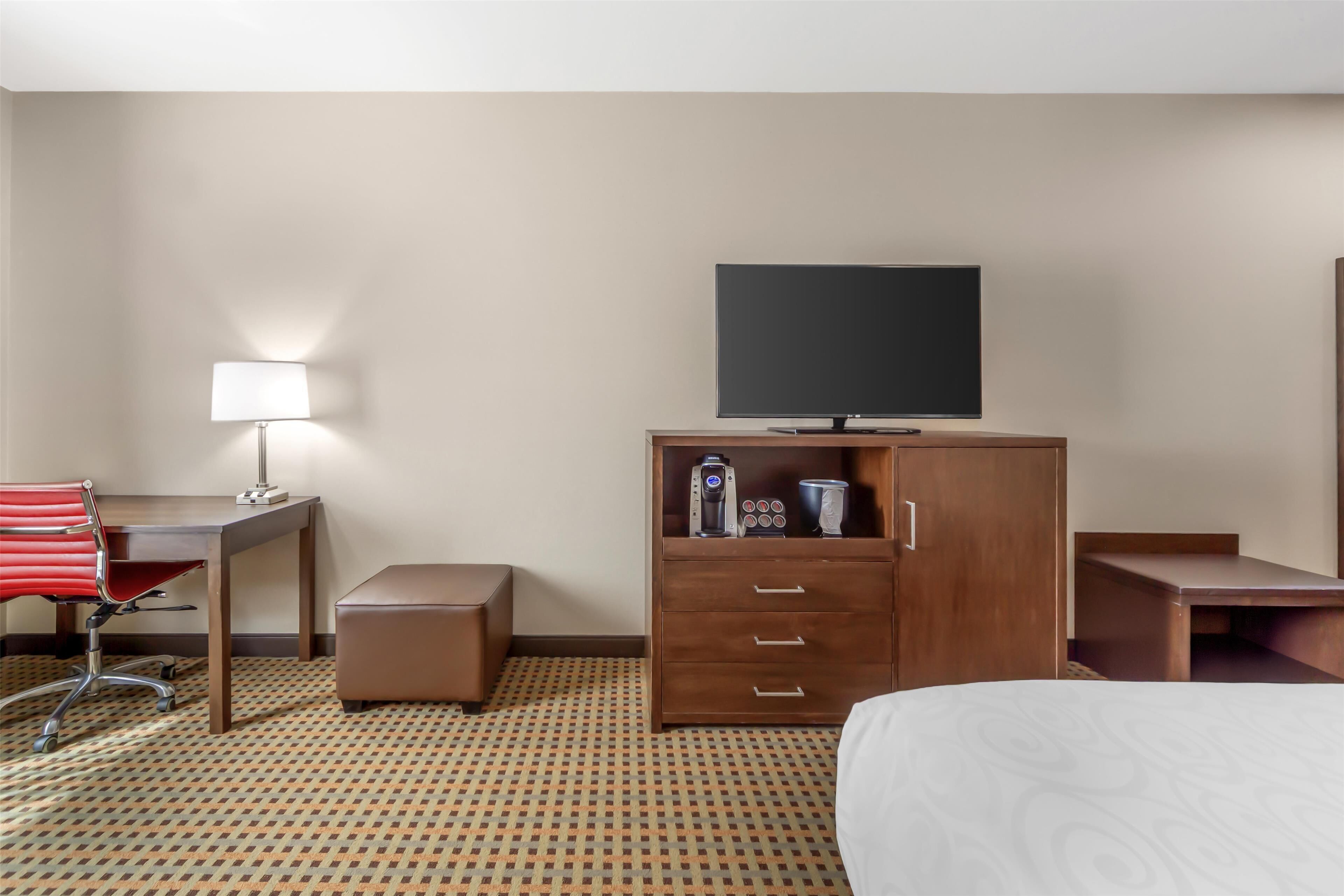 A hotel room with a bed, a desk with a chair, a wardrobe, a flat-screen TV, a coffee machine, and a lamp.