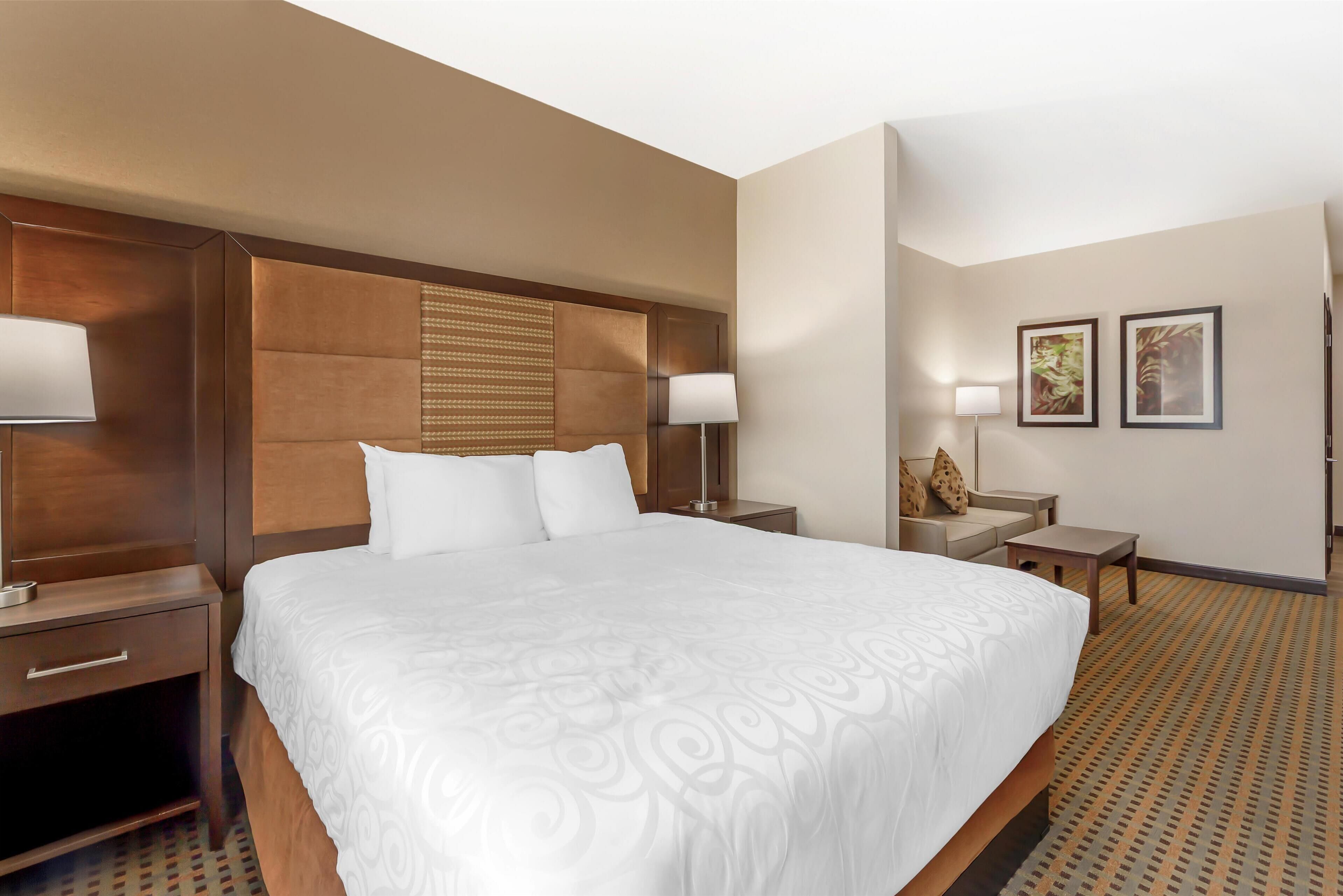 Habitación de hotel con una cama grande, cabecero de madera, mesita de noche con lámpara, una pequeña mesa con una silla y dos cuadros en la pared.