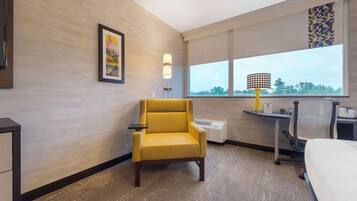 Habitación de hotel con un sillón amarillo, un escritorio con lámpara, una cama y un ventanal con vistas.