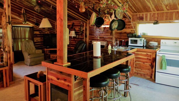 A rustic cabin interior with a wooden bar, stools, a fireplace, and a kitchen area with a stove and microwave. - Cedar Cabin Located on a Buffalo Farm Near the Buffalo National River (Marshall)