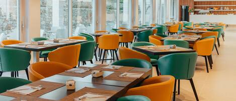 A modern restaurant with large windows, green and orange chairs, and neatly arranged tables.