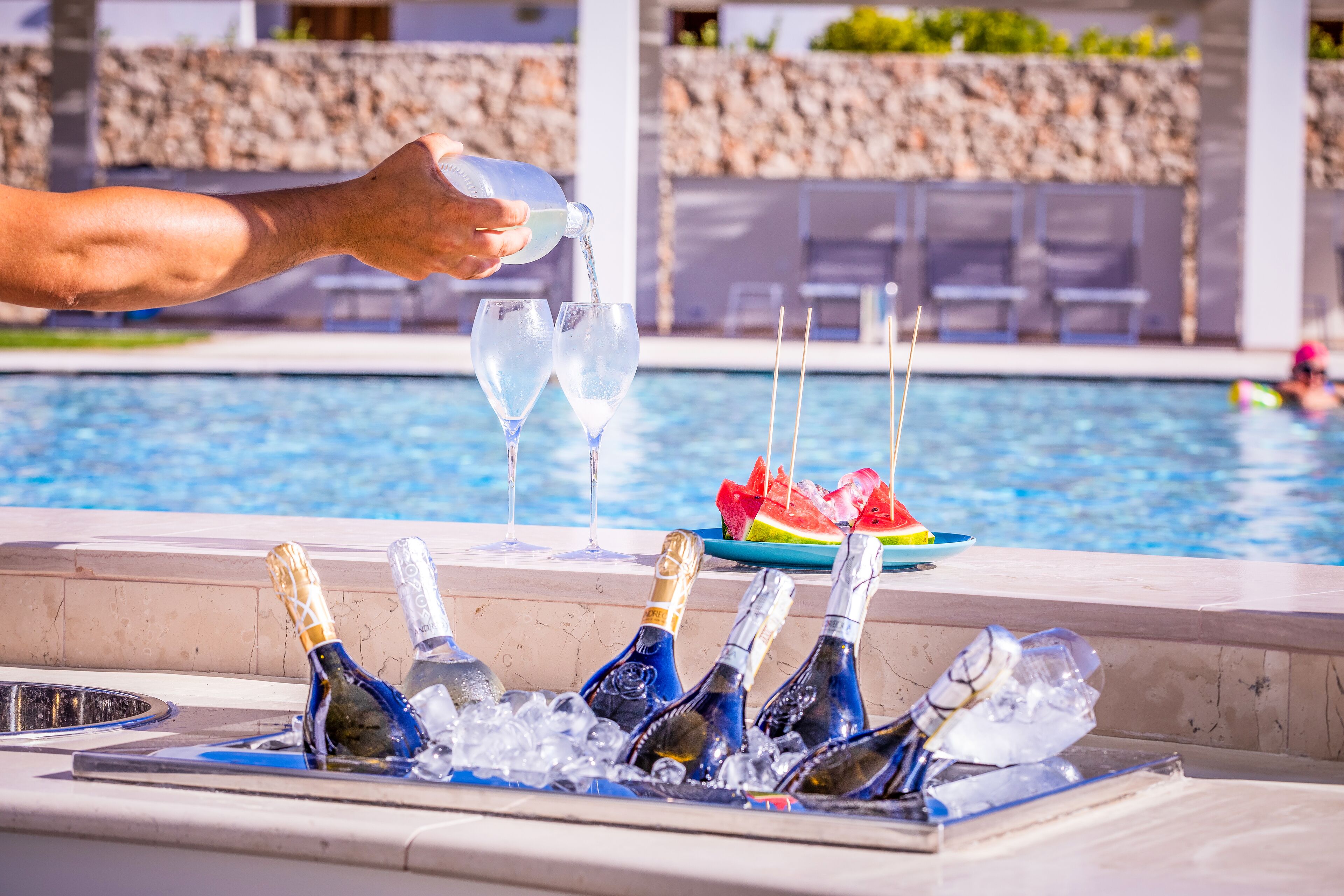 Escena junto a la piscina con una persona sirviendo una bebida en un vaso, botellas de champán en hielo y una sandía sobre una mesa.