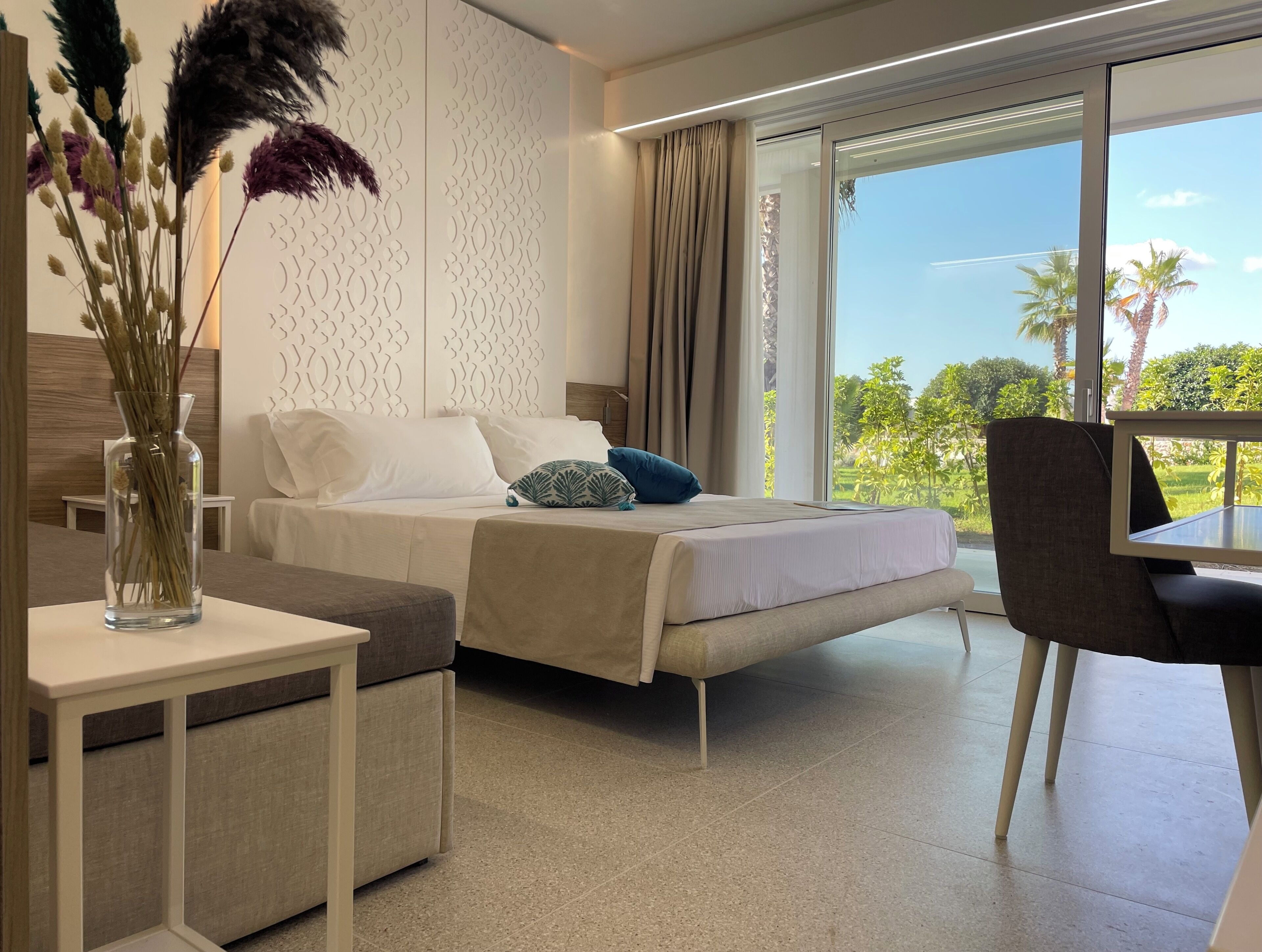 Une chambre d’hôtel moderne, équipée d’un lit, d’une table de chevet et offrant une vue sur la verdure grâce à de grandes fenêtres.
