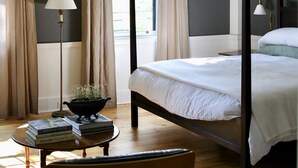 A four-poster bed with a white bedspread, a bedside table with a lamp, and a chair in a room with wooden flooring and large windows. - Inn at Kenmore Hall (Richmond)
