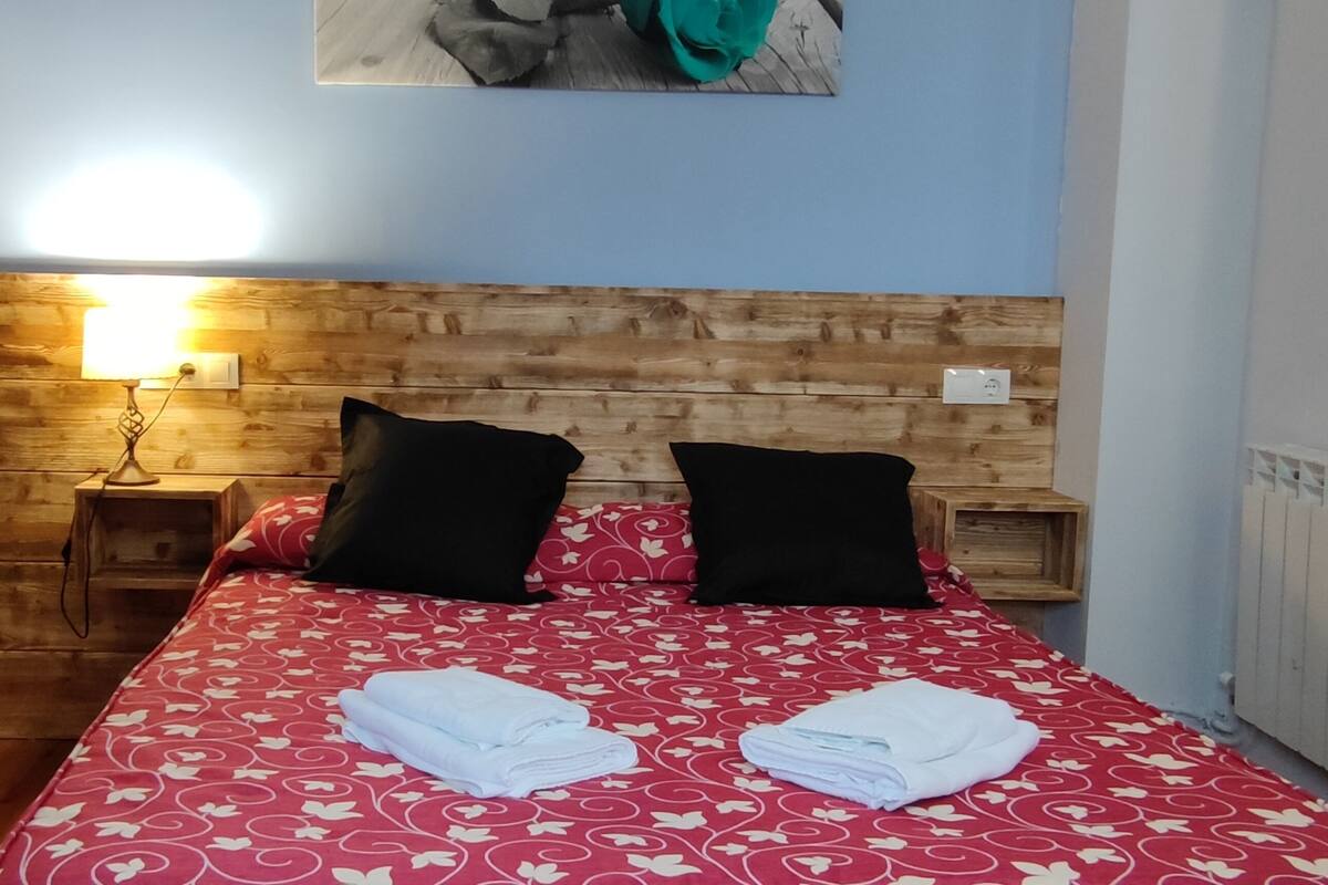 A bedroom with a wooden headboard, a red patterned bedspread, two black pillows, a bedside lamp, and a folded towel on the bed.