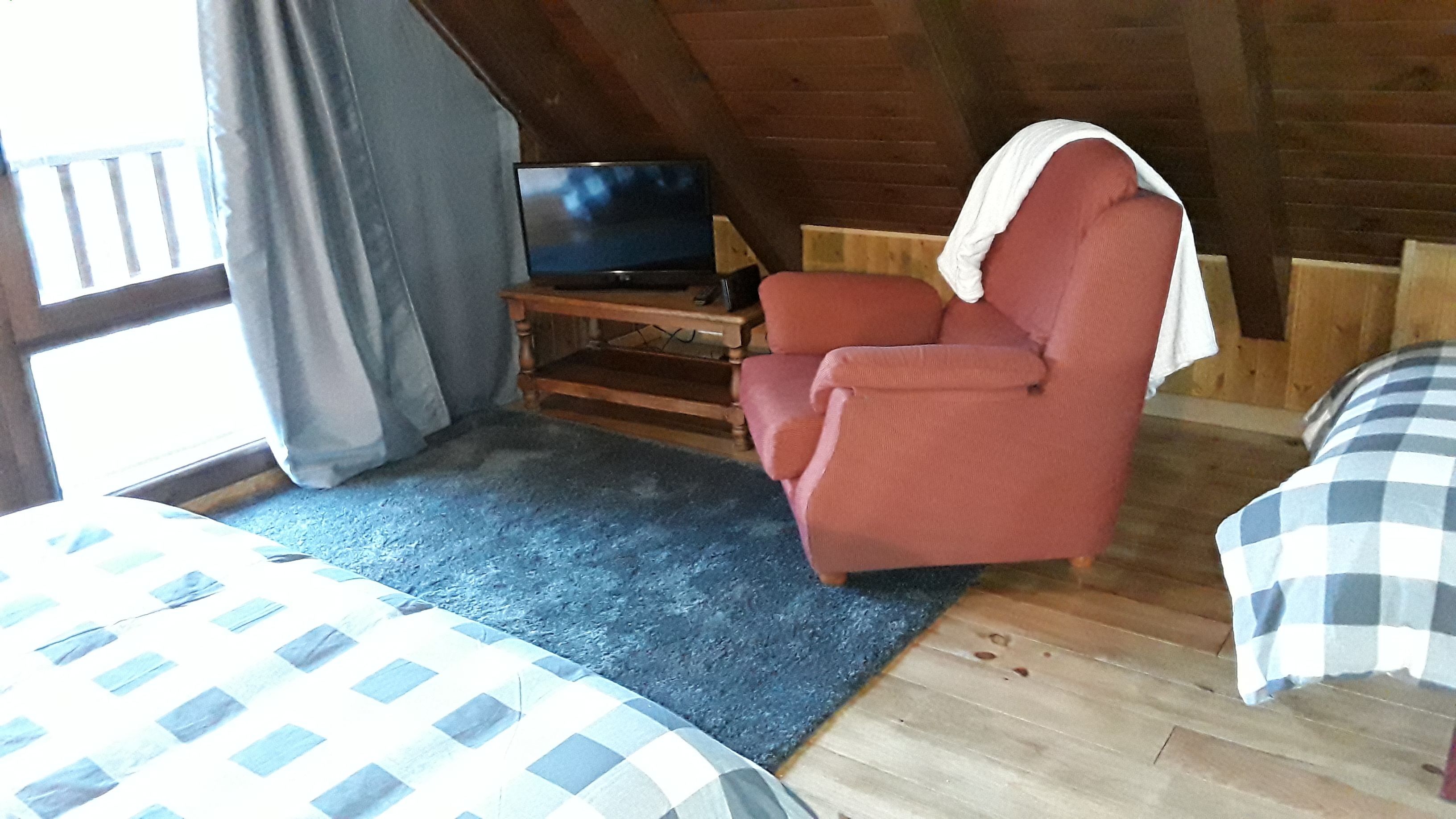 Une pièce confortable avec un lit, un fauteuil rouge, un téléviseur posé sur un meuble en bois et une fenêtre avec des rideaux.