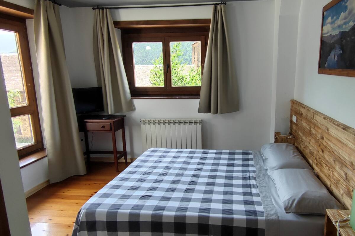 A bedroom with a checkered bedspread, a wooden headboard, a radiator, a desk, and a window with curtains.