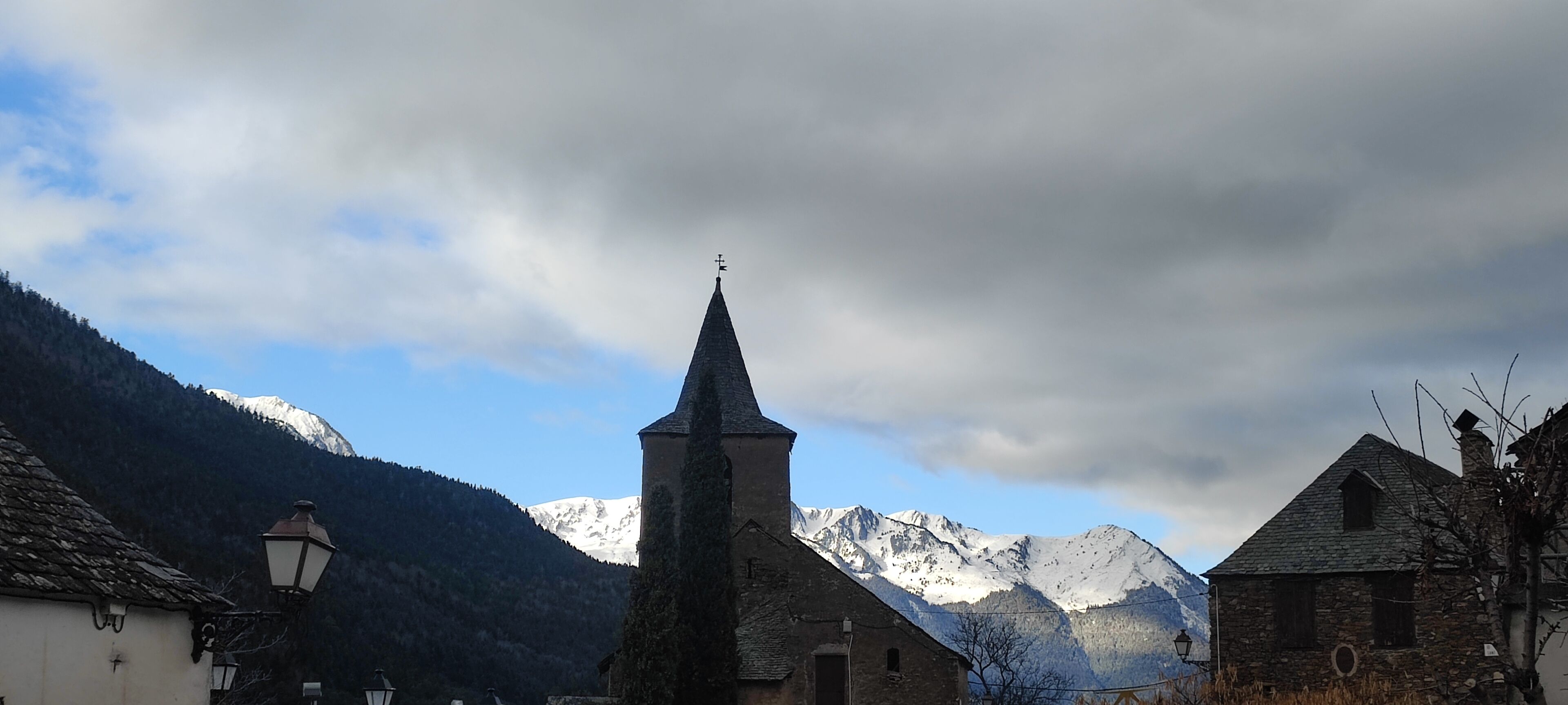 A church with a tall steeple, surrounded by mountains and a cloudy sky.