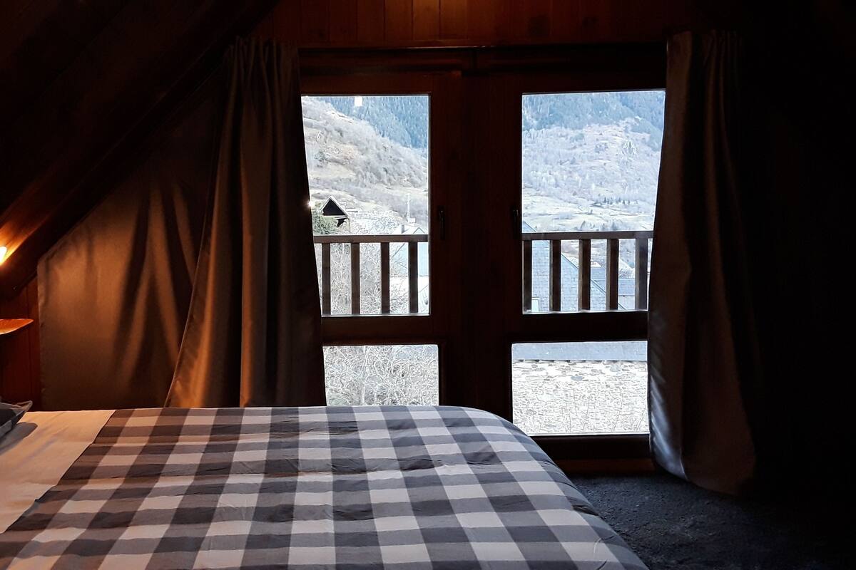 A room with a checkered bedspread, wooden headboard, and a window with a view of a snowy landscape.