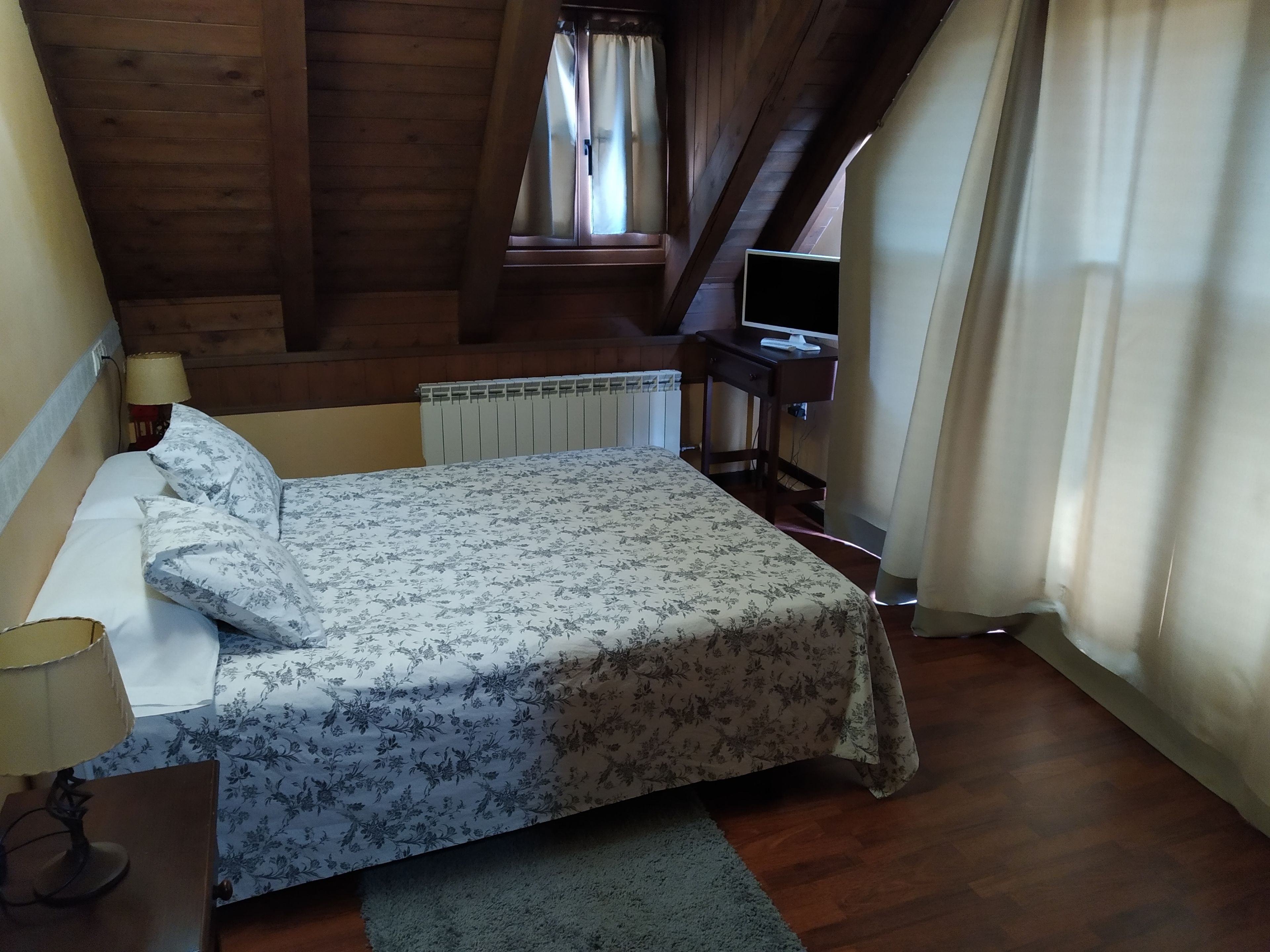 Un dormitori amb llit, escriptori, cadira, làmpada i finestra amb cortines.