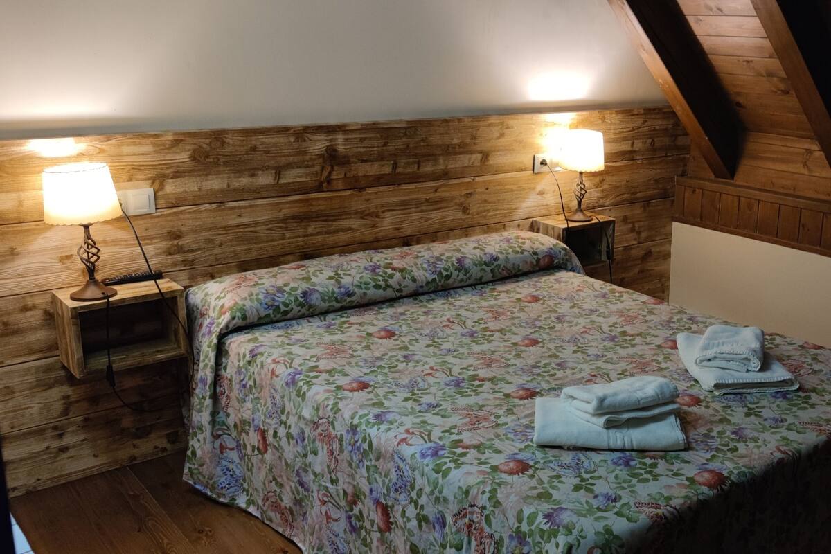 A bedroom with a wooden headboard, two bedside lamps, a floral bedspread, and a neatly made bed with folded towels.