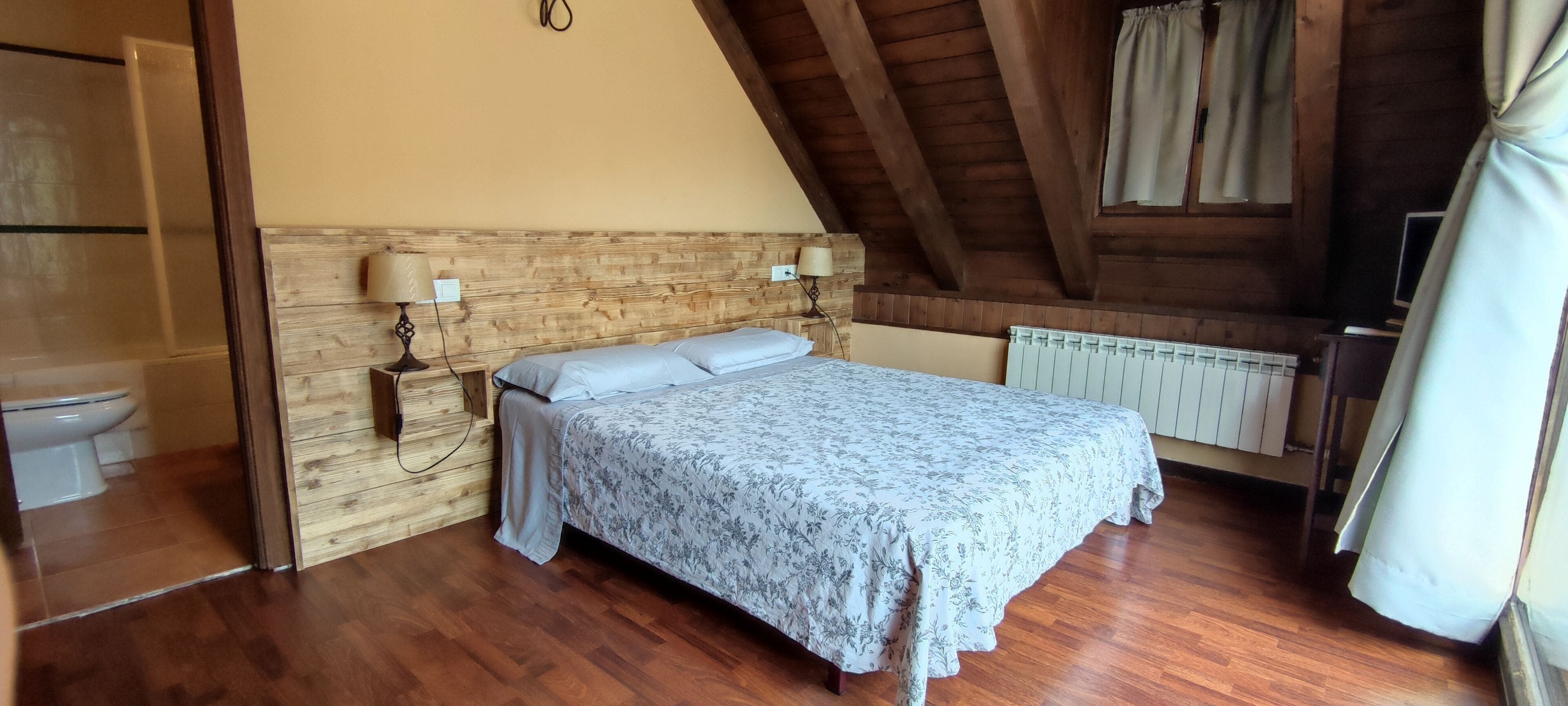 A bedroom with a wooden headboard, a bed with a floral bedspread, two bedside lamps, a radiator, and a window with curtains.