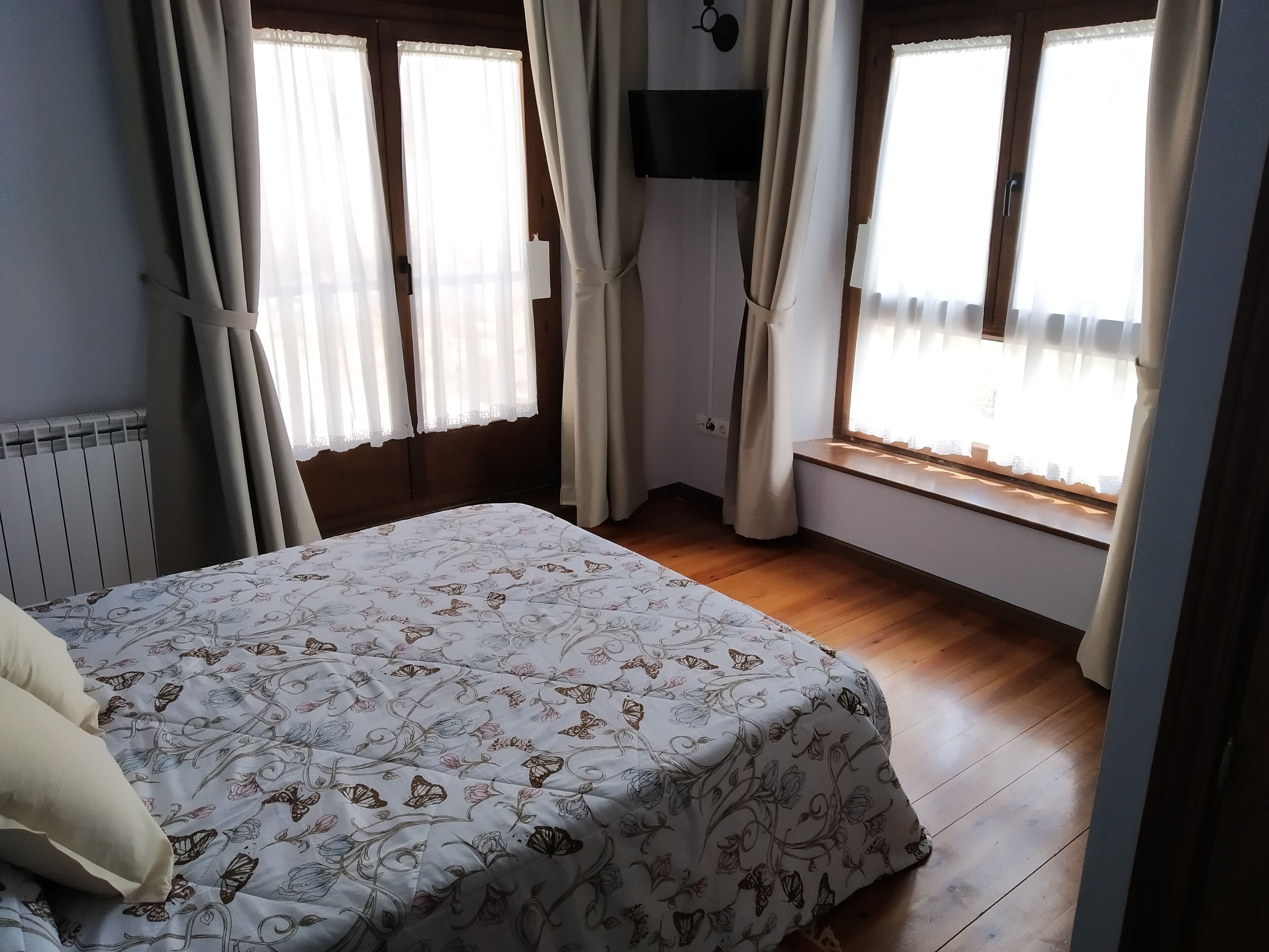 A bedroom with a bed, wooden floor, and large windows with curtains.